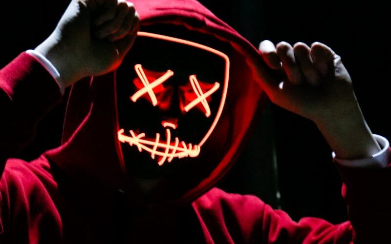 A spooky illuminated mask figure with a red hoodie in a dark setting, perfect for Halloween themes.